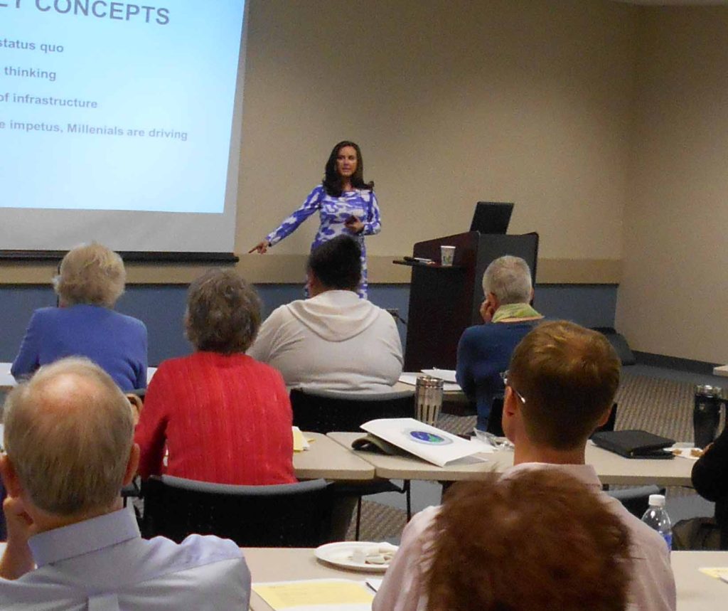 Esther Greenhouse, standing by a lectern and in front of a large screen, teaching a group of seated adults.