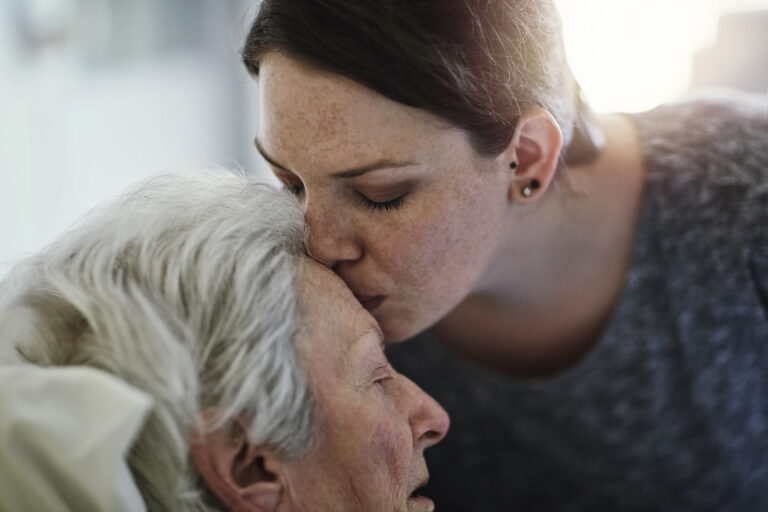 An adult daughter leaning down and kissing the forehead of her elderly mother who is in bed