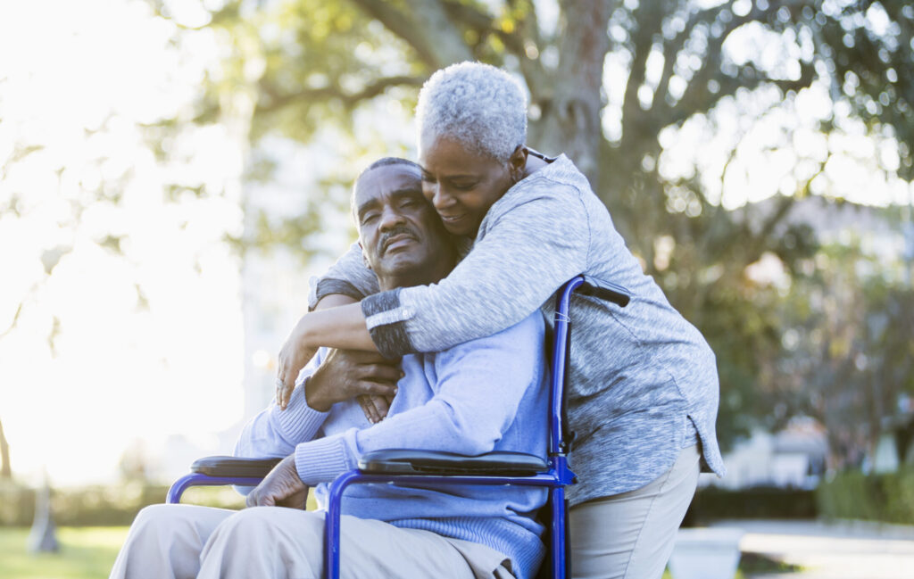 Portrait of a senior African American couple outdoors, showing their affection in the bright sunlight. The man is sitting in a wheelchair, in the warm embrace of his devoted wife. Their eyes are closed.